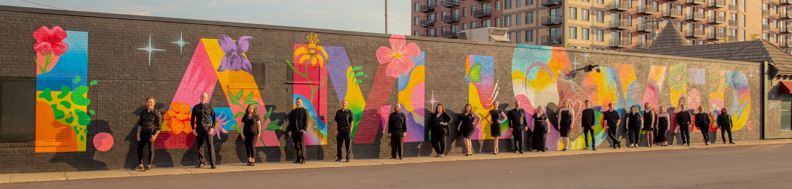 The members of KC VITAs chamber choir standing in front of a colorful mural that reads 'I' am loved'.